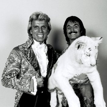 Two people in ornate jackets hold a white tiger cub against a plain backdrop.