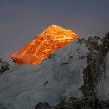 Mountain peak glows orange, illuminated by sunlight against a clear sky, surrounded by rugged snow-covered slopes.