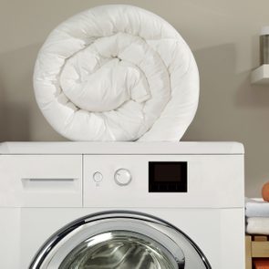 Rolled white duvet resting on a washing machine in a laundry room with shelves of neatly folded towels and bottles.
