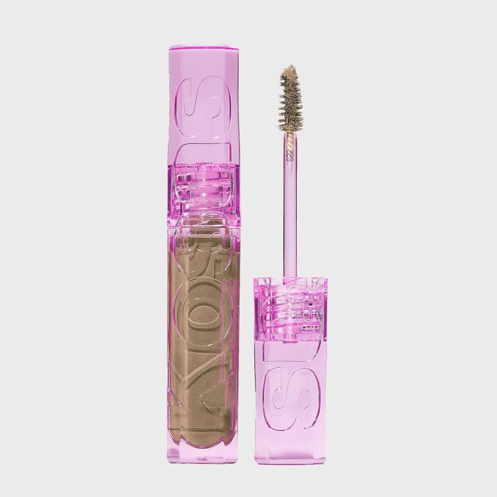 Mascara wand displays beige product, housed in a transparent pink tube with engraved letters, set against a plain white background.