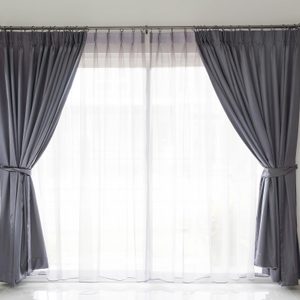 Gray curtains are tied back, revealing sheer white drapes, in a bright room with a tiled floor and a leather sofa.