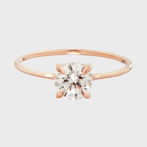 A round diamond is set in a rose gold band; simple elegance against a plain, light gray background.