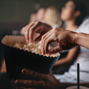 Hands reach for popcorn, sitting in a darkened movie theater amidst an absorbed audience.
