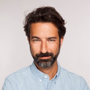 A man with a beard smiles subtly, wearing a light blue button-up shirt, set against a plain, neutral background.