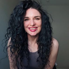 Person smiling, leaning forward with curly hair, in front of a plain wall.