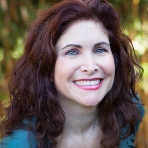 A person smiles brightly, surrounded by lush, blurred greenery in the background.