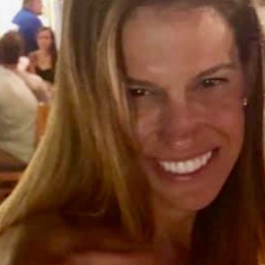 Smiling woman with long hair leans forward in a busy indoor setting, with people sitting and soft lighting overhead.