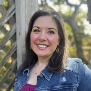 Woman smiling, wearing a denim jacket, standing outdoors next to a wooden lattice, with sunlight filtering through trees in the background.