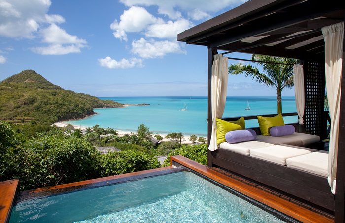 Lounge bed overlooks infinity pool and turquoise bay with sailboats, nestled in lush, tropical setting under a clear blue sky.