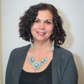 A woman smiles warmly, adorned with a large turquoise necklace and earrings, against a plain, light-colored background.