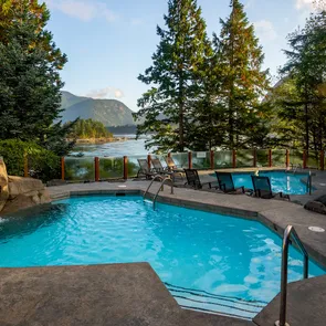 A swimming pool sits quietly surrounded by trees and mountains, with lounge chairs arranged nearby under a clear sky.