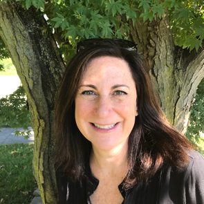 A person smiles beneath a large tree, with textured bark and green leaves, in a sunlit garden setting.