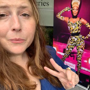 Person making a peace sign gesture while posing near a vibrant poster of a performer in a colorful outfit.
