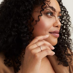 A woman with curly hair holds her chin, showcasing delicate rings. She is wearing a tan outfit, set against a neutral background.