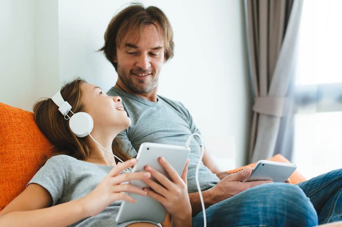 Father and pre teen daughter playing on tablet together on couch in room at home. Good parent and child relations.