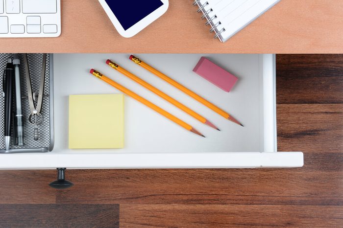 Top view of an open desk drawer showing the items inside. The top of the desk has a computer keyboard Cell Phone and note pad. The neat drawer has pencils paper and organizer.