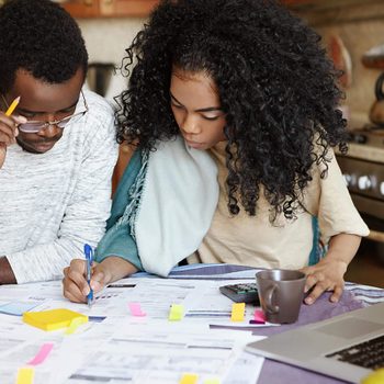 Young confident African housewife with Afro hairstyle helping her husband to manage domestic finances, calculating and making notes with pen, both sitting at kitchen table with laptop and papers