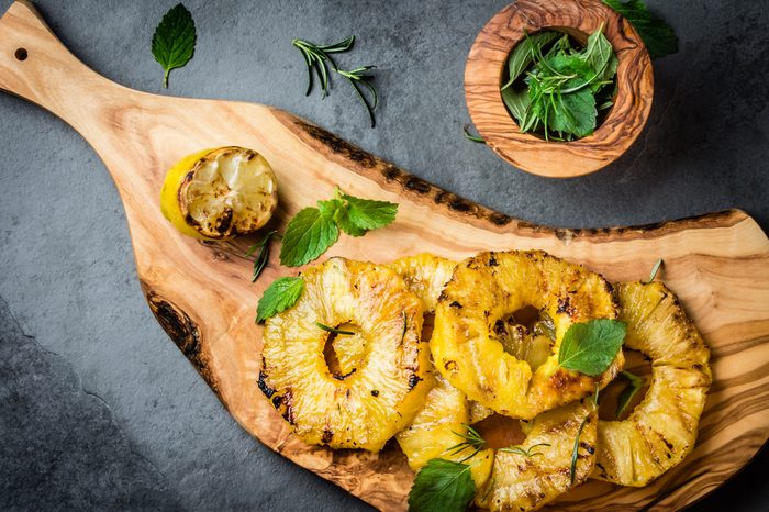 Grilled pineapple slices with fresh mint on olive wooden cutting board, gray slate background. top view