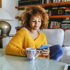 Woman in yellow sweater uses smartphone while seated on a sofa, with a cartoon mug on the table in a book-filled room.