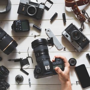 Man hand Holds Digital Camera On Table Next To Lenses And Accessories On White Wooden Background. Top View