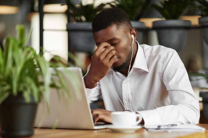 Bored, sad african man in white shirt, thinking, decided problem, working at cafe with laptop. Student male with closed eyes, holding head by hand. Freelancer have coffee break, listening music.