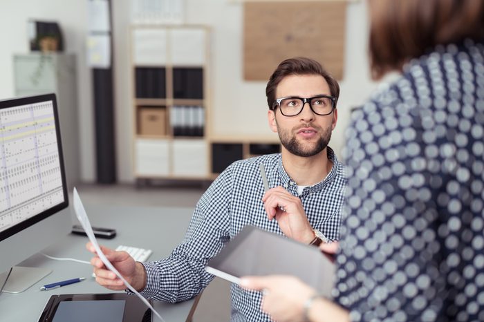 Young Handsome Office Man Talking to his Female Manager While Holding a Paper at his Working Table with Computer.