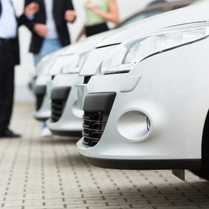 Two men man and one woman stand behind a row of silver-metallic cars on a parking place on yard of dealer