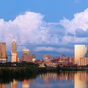 Skyscrapers reflect light against large clouds, lining a riverside cityscape.