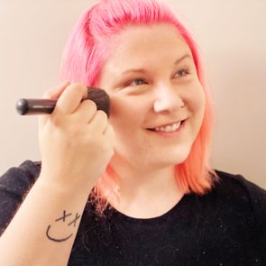 Person uses makeup brush, smiling with pink hair, wearing a black shirt indoors. Smiley face tattoo on forearm.