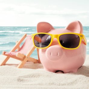 A piggy bank wearing yellow sunglasses sits on sandy beach with a striped deck chair beside it, ocean waves in the background.
