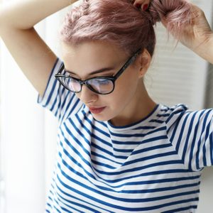 Morning routine. Beautiful young female with pinkish hair wearing blue and white striped top and trendy eyeglasses standing at window and doing hairstyle, going out to have coffee with friends