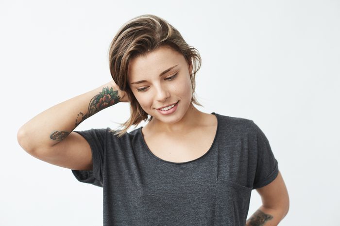 Shy young pretty tattooed girl smiling looking down over white background.
