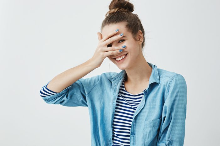 Girl saying silly words and gets embarrassed. Indoor shot of cute slender woman in stylish garment, covering eyes with hand but peeking, smiling broadly at camera, expressing positive emotions