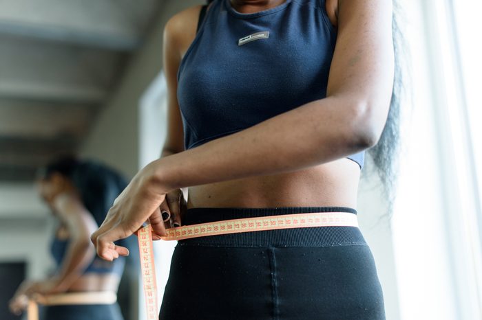 Close-up of young adult black woman in sports clothing. African american girl measuring waist with yellow tape close up