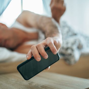 A person reaches for a smartphone from bed, under a soft blanket, in a gentle morning light.
