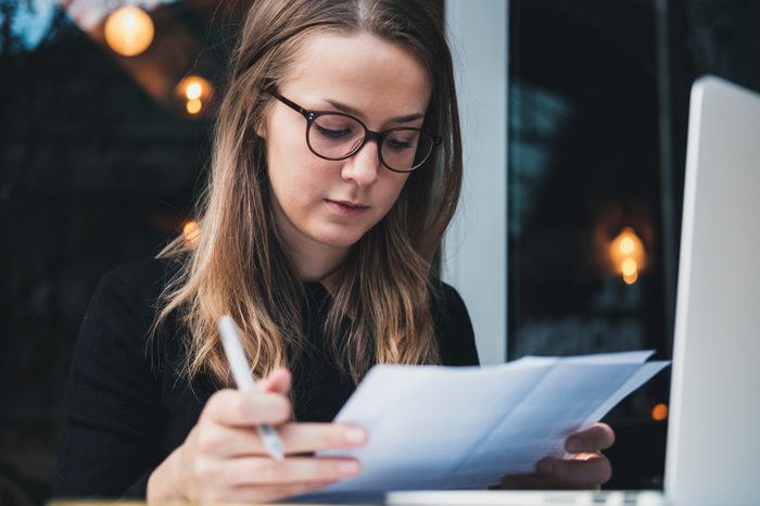 Portrait of a young beautiful girl sitting at table with laptop, holding pen in hand and reading piece of paper. Education concept