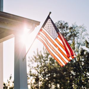 An American flag waves from a house porch, illuminated by sunlight, with trees in the background.