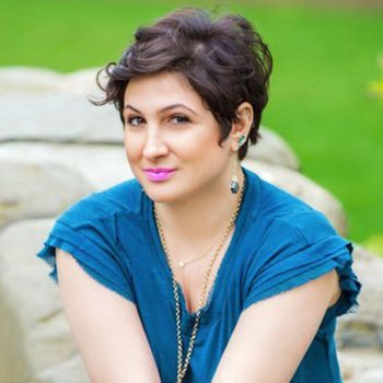 A person with short hair smiles, sitting against a stone wall, wearing a blue shirt; greenery fills the background.