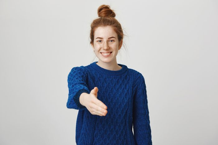 Happy to finally meet you. Studio shot of confident relaxed and friendly young woman with red hair combed in bun pulling head towards camera for handshake with cute smile, standing over gray wall