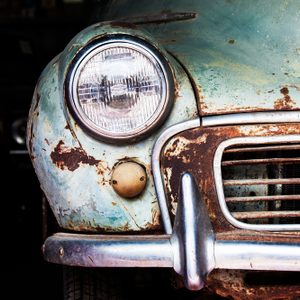 Rusty car shows aged front view with headlight and grille, positioned in a dimly lit garage setting.