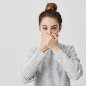 A person covers their mouth with both hands, wearing a gray sweater, set against a plain white background.