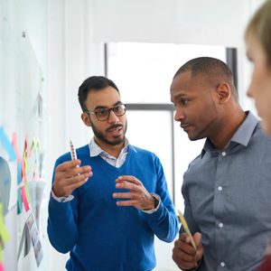 Three people collaborate at a whiteboard covered in colorful sticky notes in a bright office, discussing ideas actively.