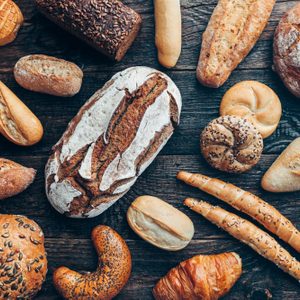 Various types of bread are arranged on a rustic wooden table, showcasing different shapes, sizes, and textures in a bakery-like setting.