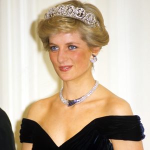 Woman wearing a tiara smiles, adorned with a necklace and earrings, in a formal indoor setting.