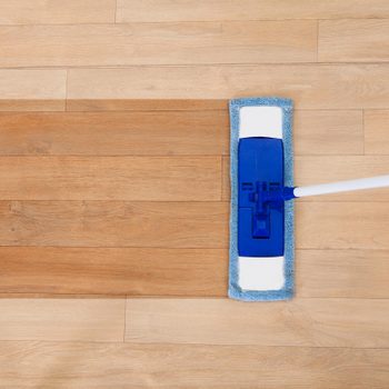 A blue mop scrubs a wooden floor, creating a clean streak across the brown planks.