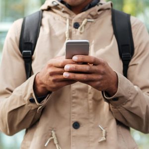 A person holds a smartphone, engaged in typing, wearing a beige jacket with a backpack, outdoors with a blurred leafy background.