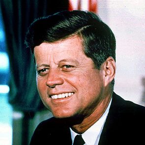 Smiling man gazes sideways, wearing a suit and tie, with curtains in the blurred background.