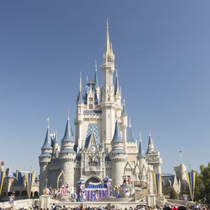 Castle stands prominently; characters perform on stage; surrounded by audience, under clear blue sky and trees.