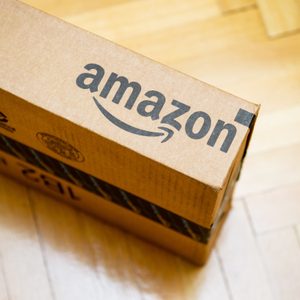 A cardboard Amazon package sits unopened on a wooden floor, displaying company logo clearly.