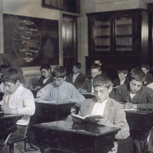 Students attentively read and write at desks in a classroom. A teacher assists a student; chalkboard diagrams are visible in the background.